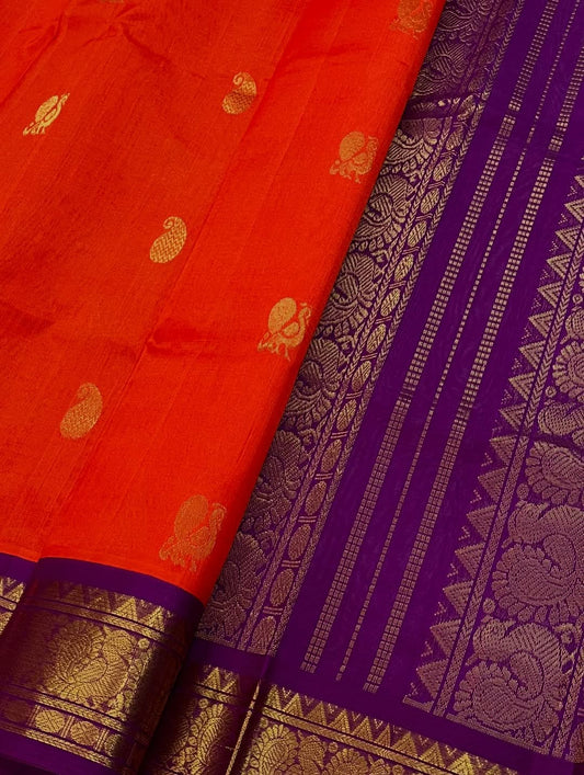 An exquisite blend of tradition and vibrancy, this silk cotton saree features a radiant orange body adorned with elegant gold zari buttas, The rich contrast border and pallu in royal purple and intricately woven with traditional gold zari motif