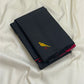 Folded black Pure Kanchipuram Silk Saree on white fabric, with a small yellow bird motif and pink edge visible.