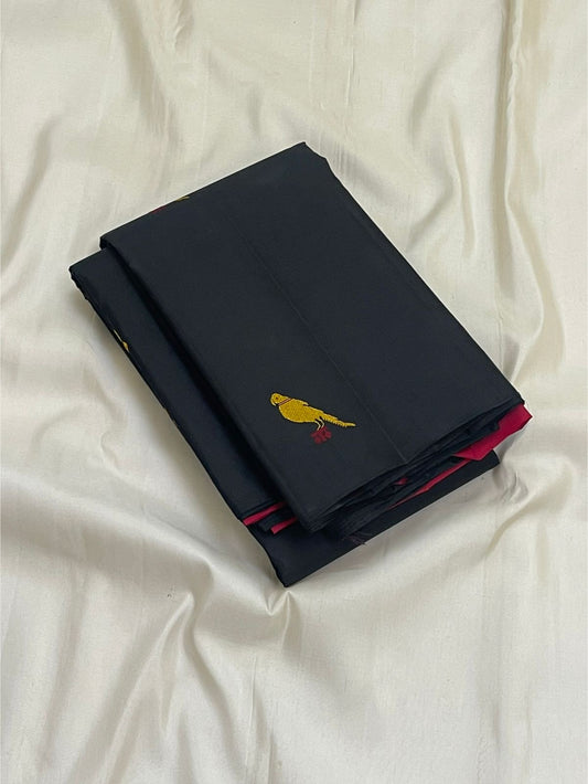 Folded black Pure Kanchipuram Silk Saree on white fabric, with a small yellow bird motif and pink edge visible.