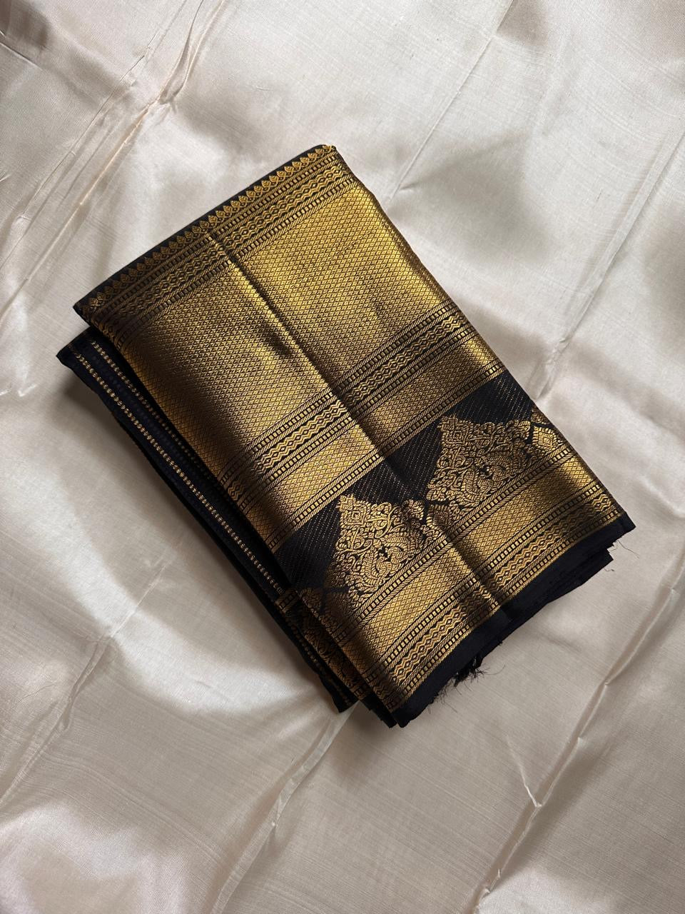 Pure Kanchipuram silk saree folded on ivory background with black body and thin gold zari stripes plus wide gold zari border