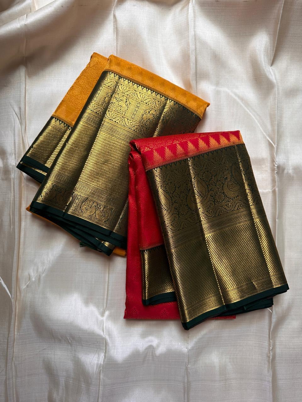 Two folded sarees shown together, one in mustard and one in red, each with a wide gold zari woven border and green edging.
