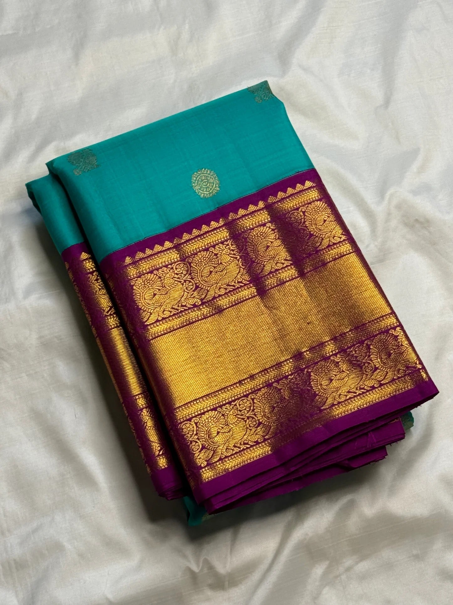 Folded Turquoise Blue Kanchipuram Korvai saree showing Deep Purple border with broad antique-gold zari and figurative motifs.