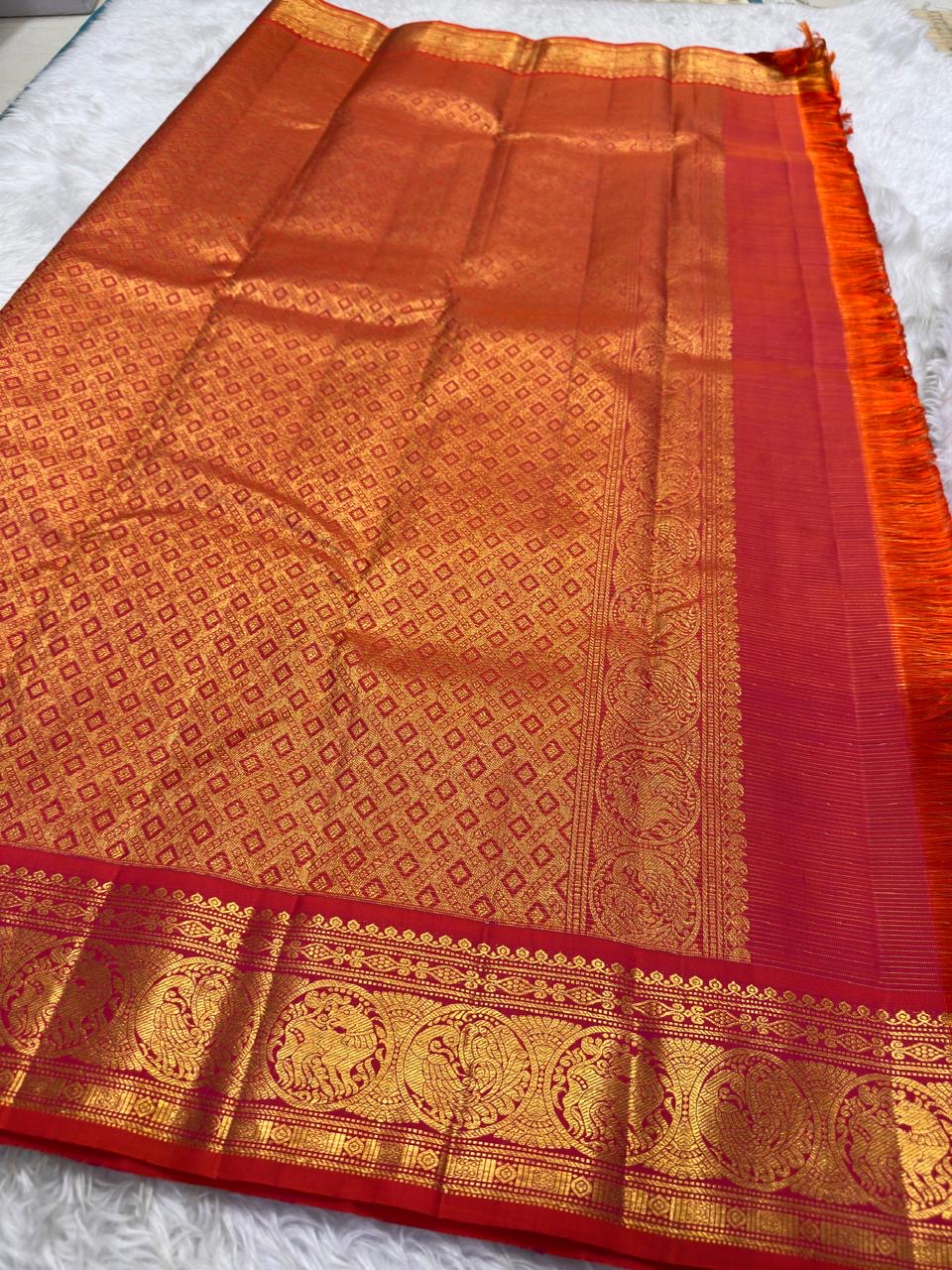 Tomato red pink shot colour Kancheepuram silk saree folded showing Vairaoosi body with Kamalam butta and ornate self-colour Mayil gold zari border