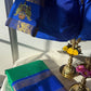 Blue saree with gold border on a white surface, next to green saree and decorative items.