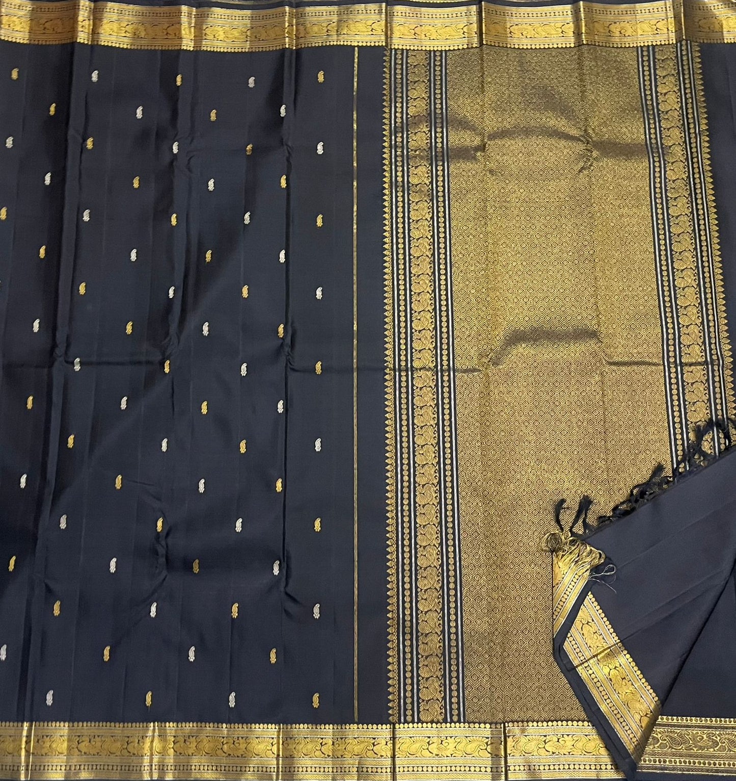 Draped view of midnight black Kanchipuram silk saree highlighting small contrast buttas and wide antique gold zari border with tassels.