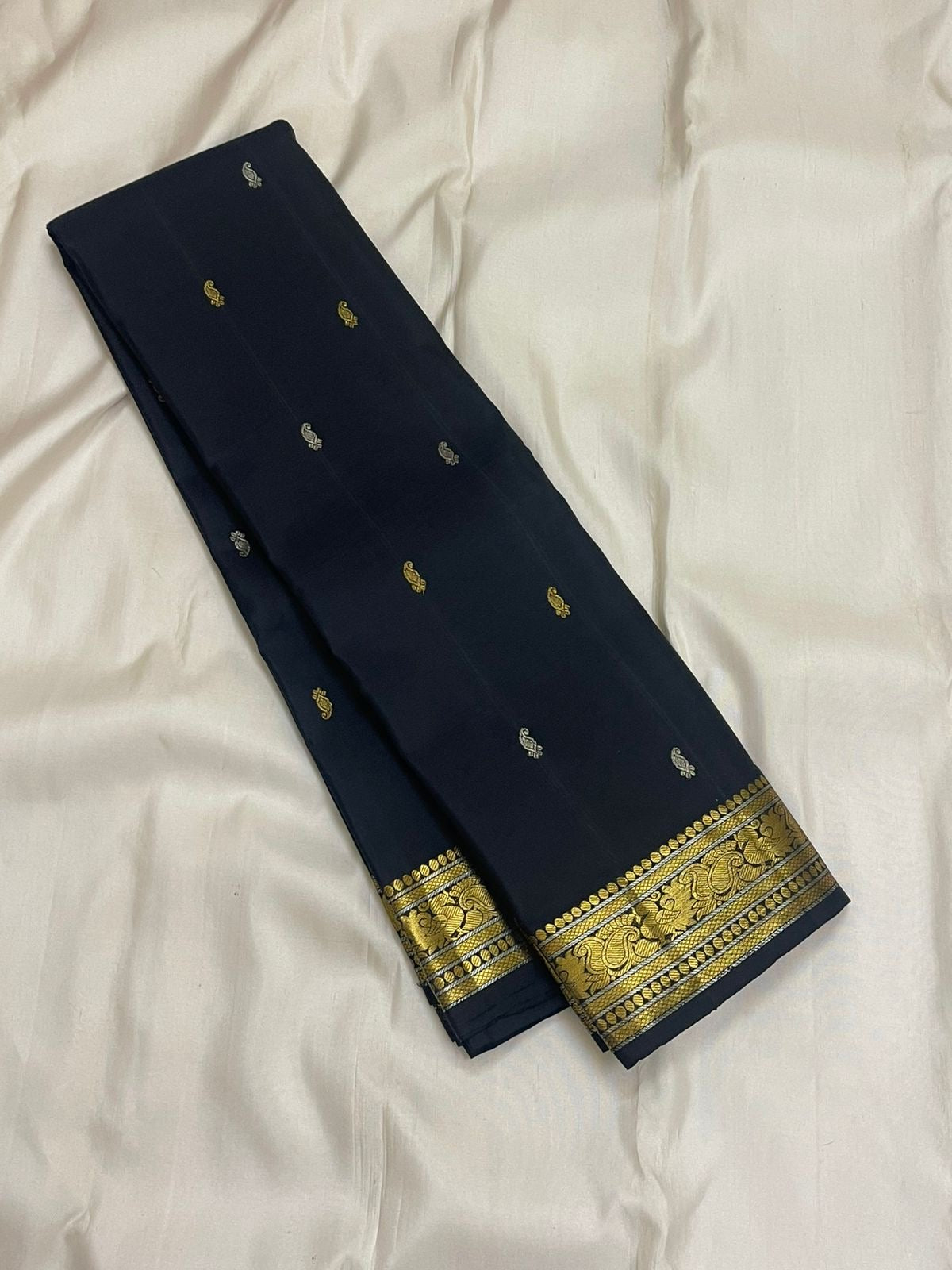 Midnight black Kanchipuram silk saree folded, featuring small contrast buttas across the body and a broad paisley border woven in antique gold zari.