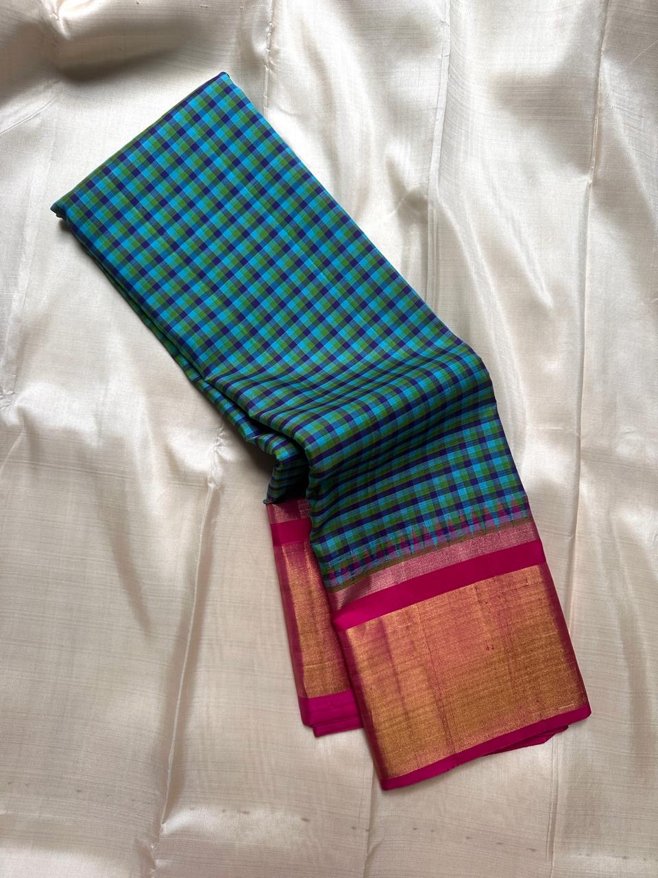 Draped blue-green checked Kanchipuram silk saree showing magenta border and wide gold zari panel on an ivory backdrop