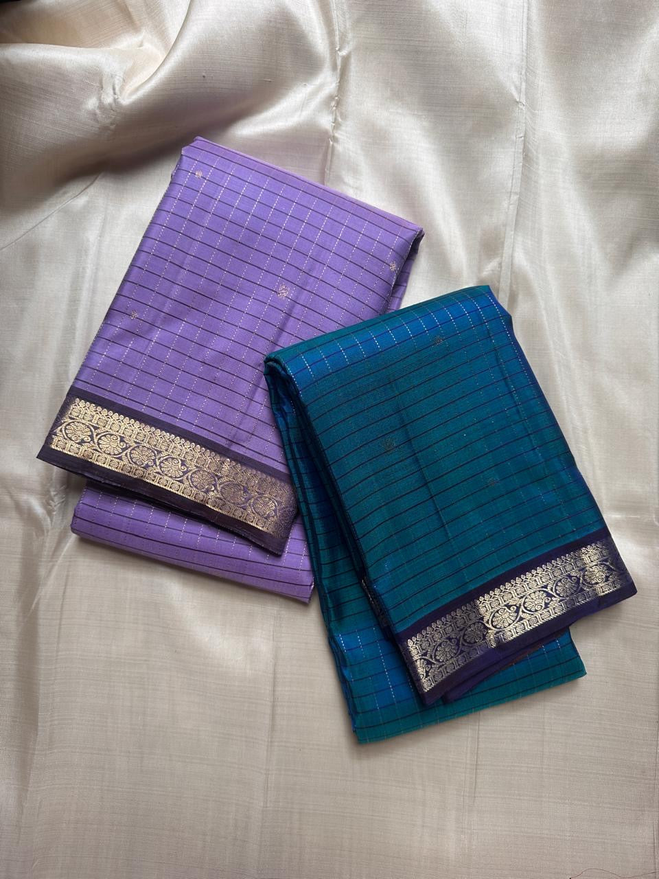 Two folded sarees on ivory fabric, one blue and one lavender, both with zari woven borders