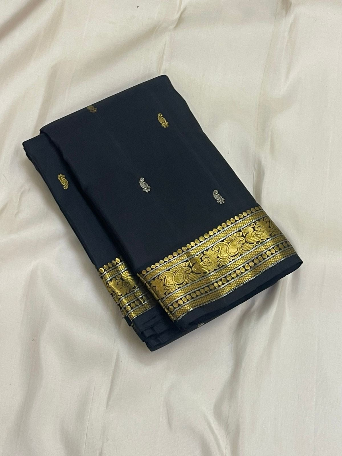 Midnight black Kanchipuram silk saree partially opened, showing evenly spaced contrast buttas and a detailed paisley border in antique gold zari.