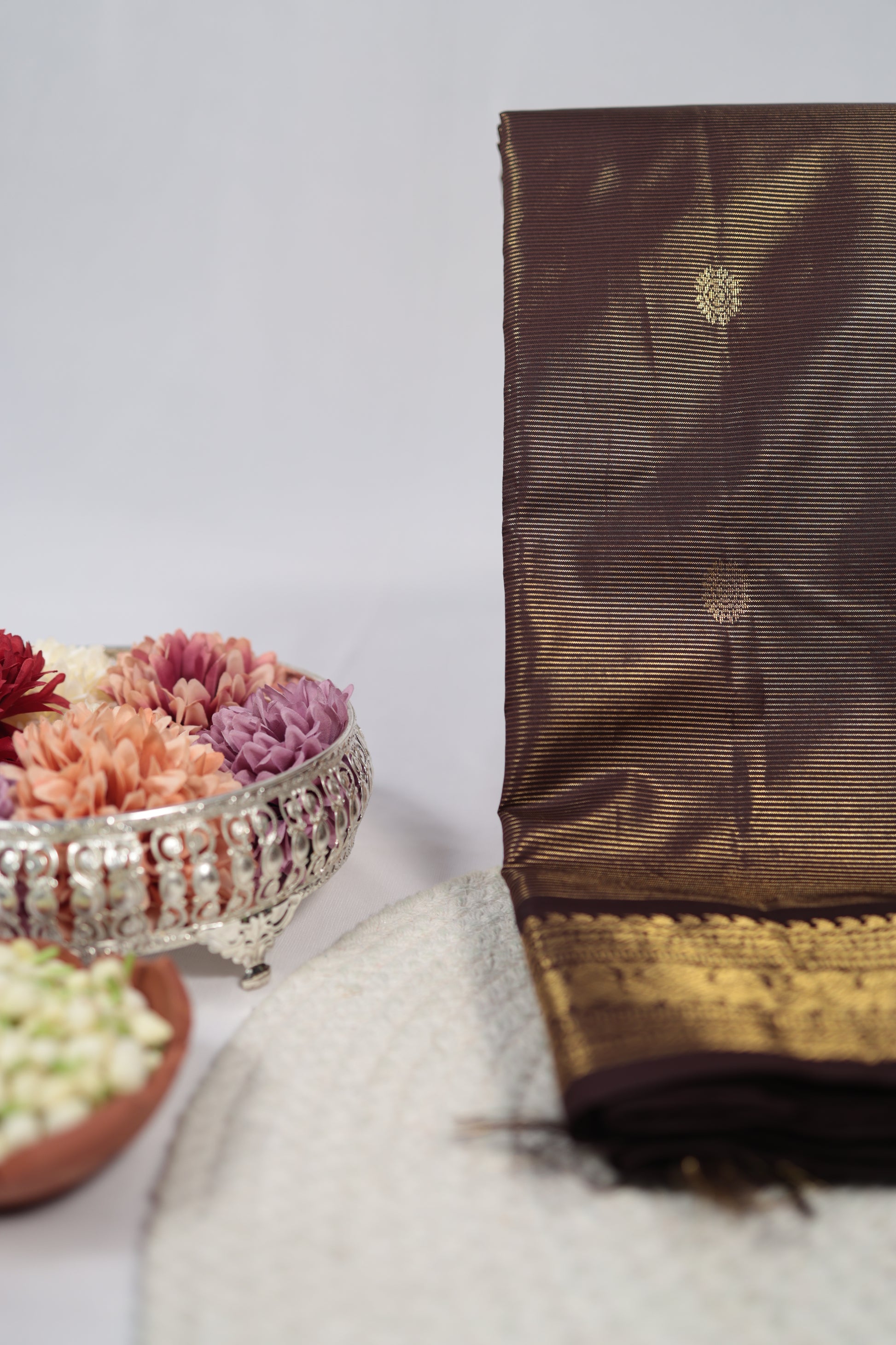 Folded deep brown vairaoosi Kanchipuram silk saree with zari woven border beside flowers and plants