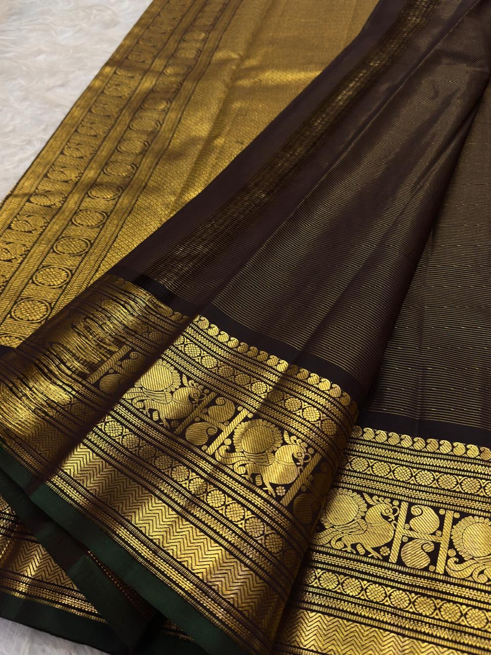 Detail of the saree border: A broad dark brown and antique-gold border with bold traditional figurative and geometric zari registers, finished with a narrow green edge.
