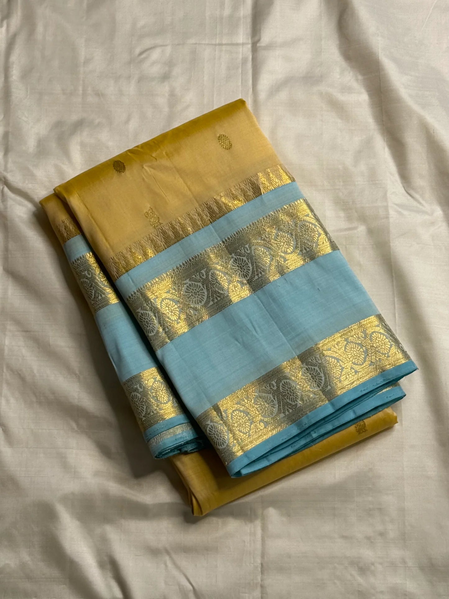 Folded butter yellow Pure Kanchipuram silk saree with sky blue border and zari woven floral band on white backdrop
