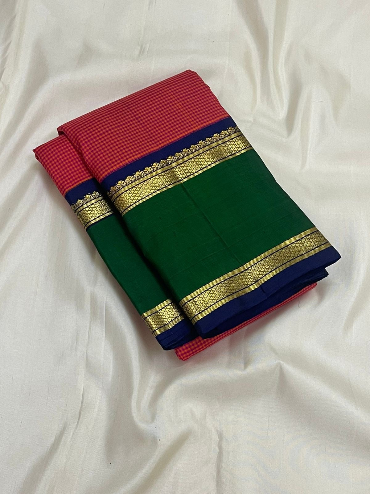 Folded pure Kanchipuram silk saree with red checks, green panel, navy border, and gold zari bands on white fabric backdrop.