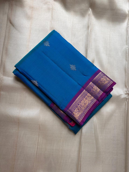 Folded teal blue Pure Kanchipuram silk saree with small zari buttas and a purple zari woven border with gold detailing.