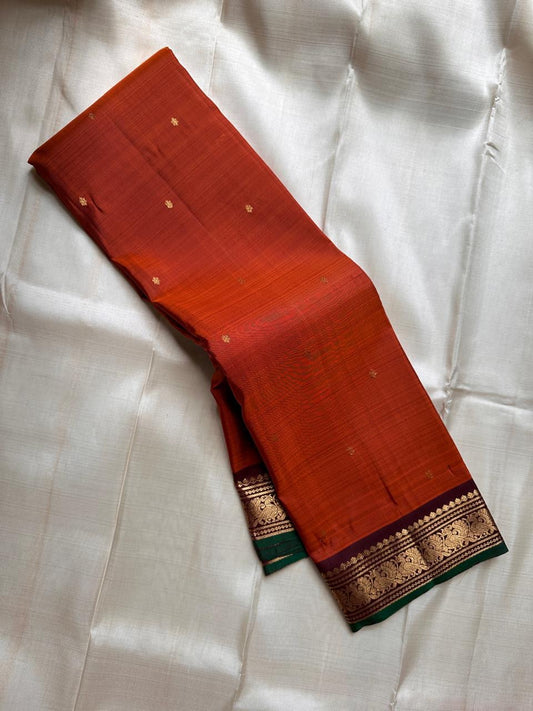 Folded rust orange Pure Kanchipuram silk saree showing small zari buttas and a maroon zari woven border with green contrast edging.