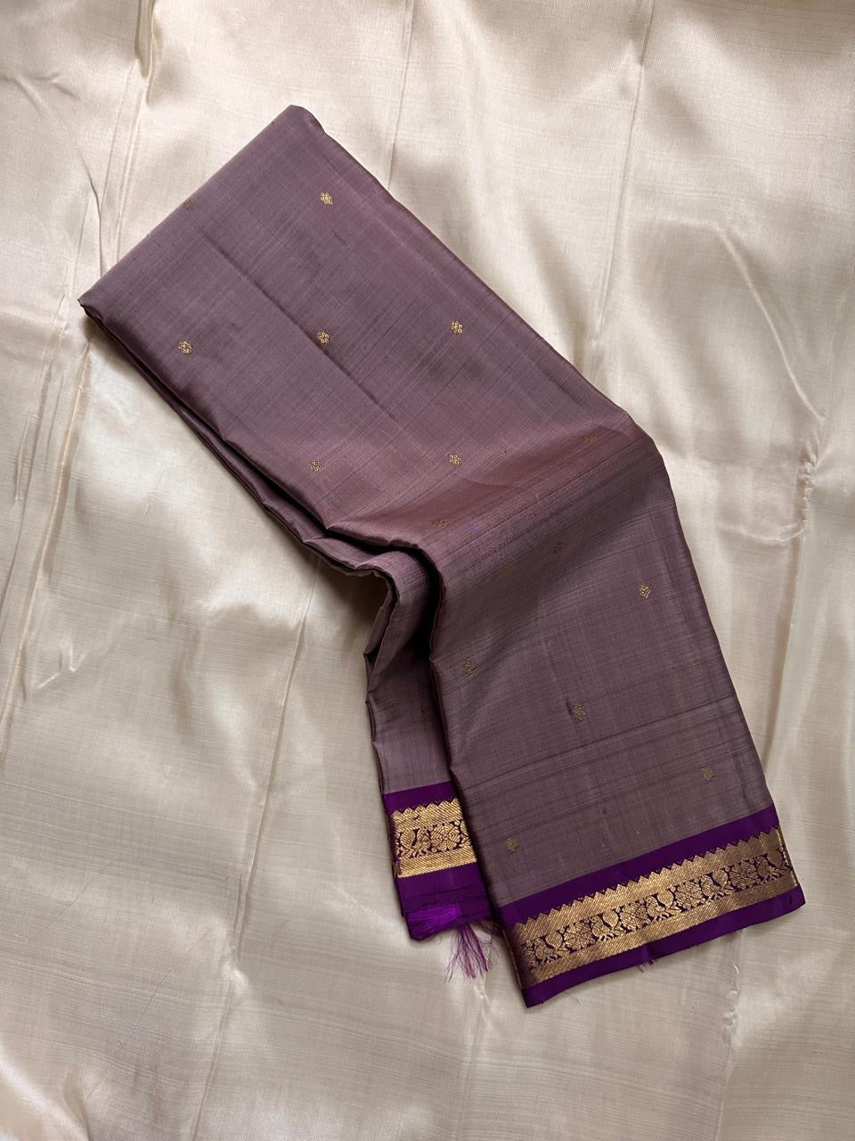 Angled fold of mauve Kanchipuram silk saree showing scattered gold buttas and a purple zari woven border edge.