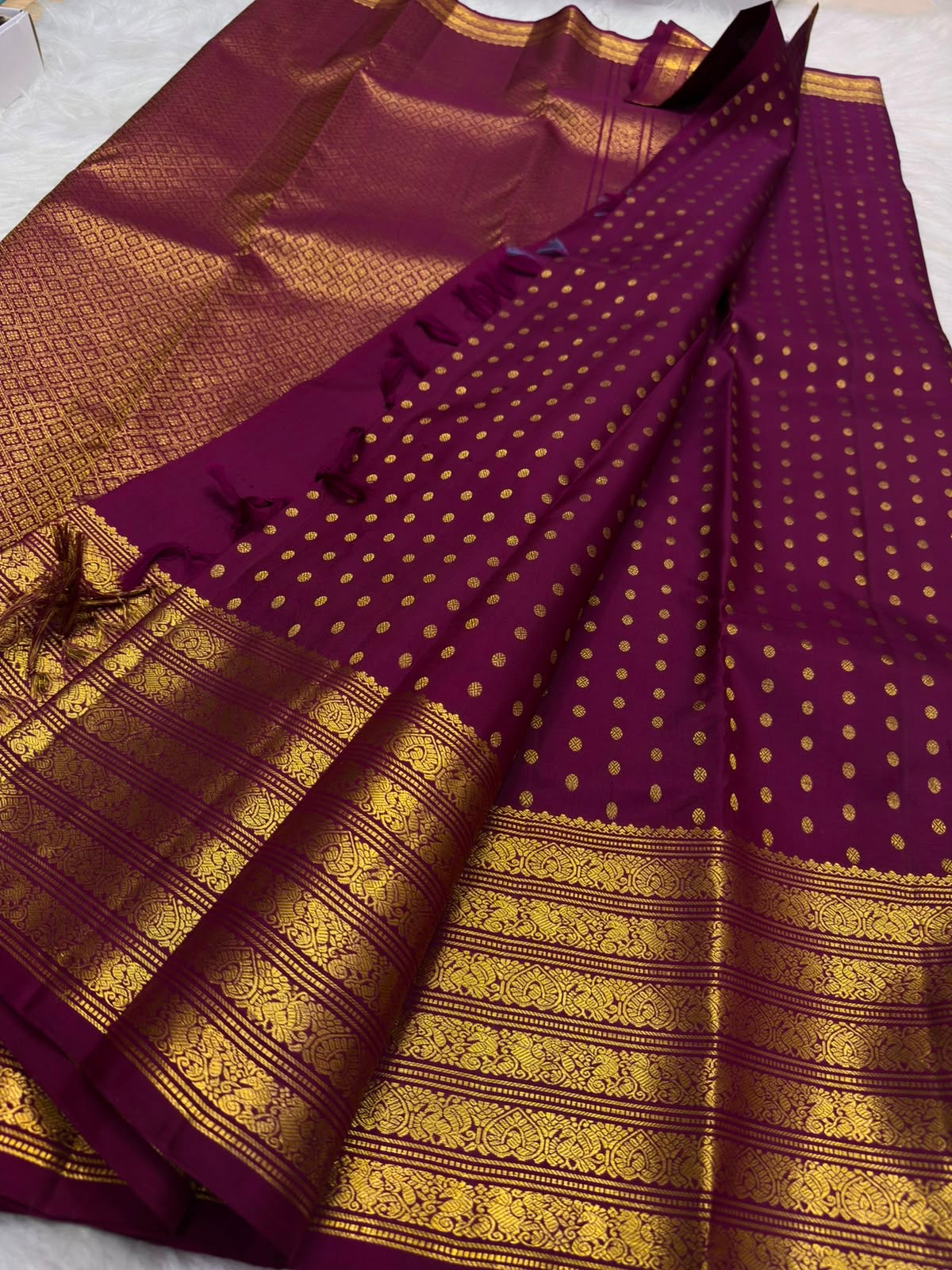 Close view of small circular gold zari buttas arranged across a deep cherry red-purple Kanchipuram silk body.