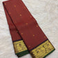 Dark plum-toned border with gold peacock and malar kodi floral vine zari and deep-green selvedge on vermilion red Kanchipuram silk saree