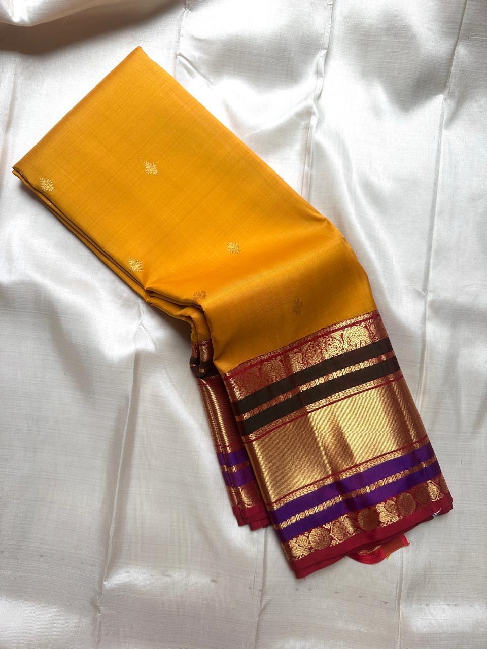 Mustard yellow Pure Kanchipuram Silk Saree with small gold buttas and wide zari border with maroon and purple accents.