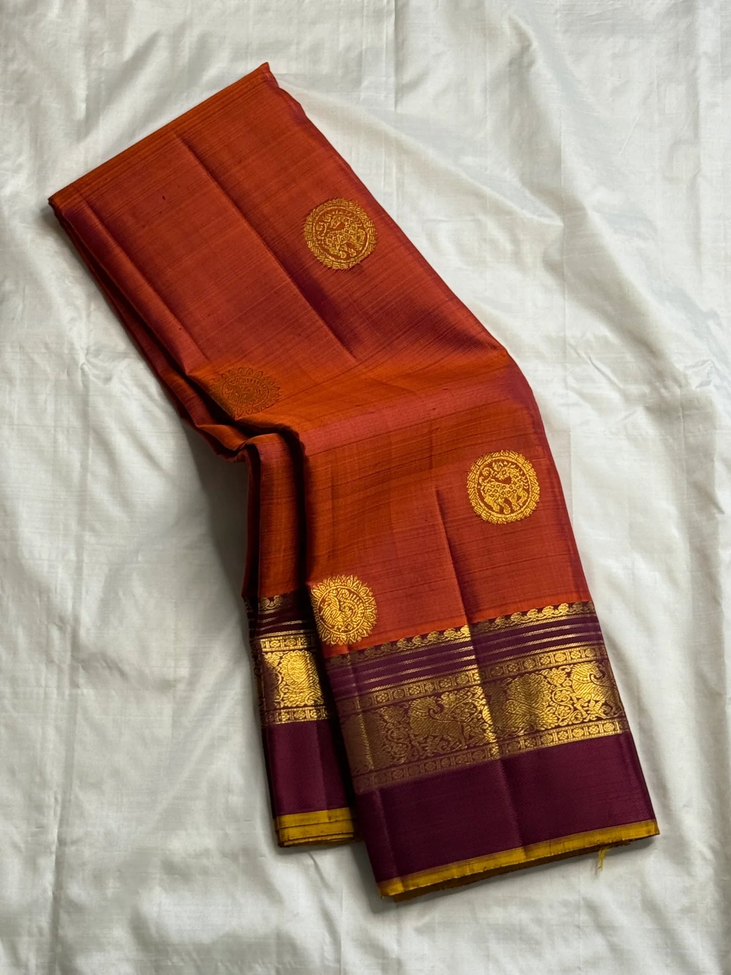 Folded rust orange Pure Kanchipuram silk saree with deep purple border and gold zari woven detailing