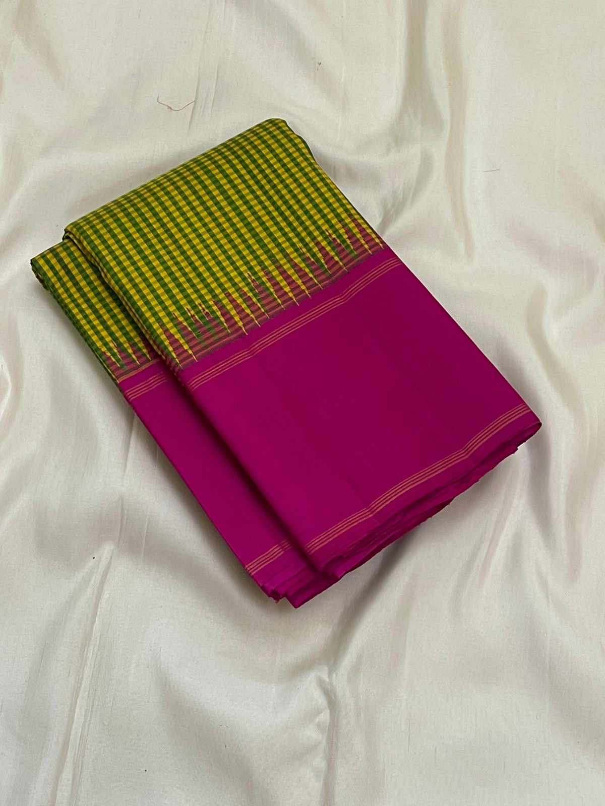 Folded pure Kanchipuram silk saree with green-yellow checks and solid magenta section with thin border lines on white backdrop