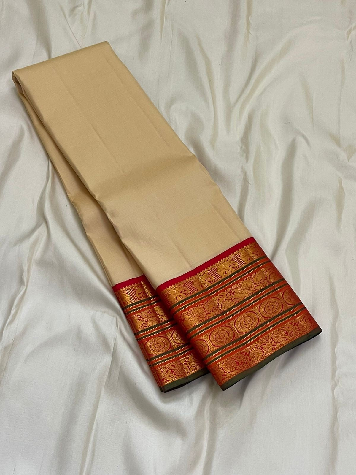 Folded ivory pure Kanchipuram silk saree with red zari woven border, photographed on a white sheet backdrop.