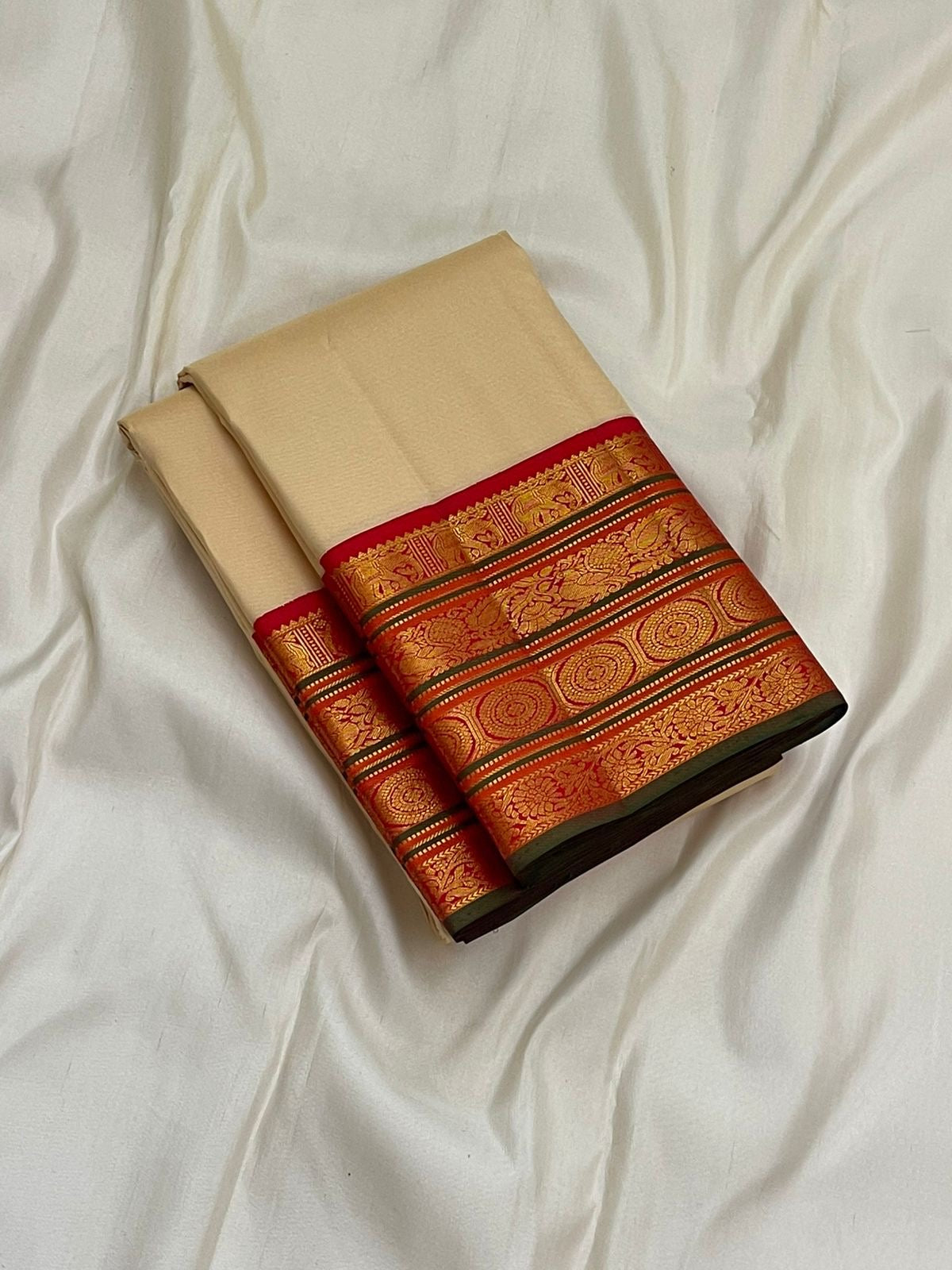 Folded pure Kanchipuram silk saree in ivory with red zari woven border, shown on white fabric background.