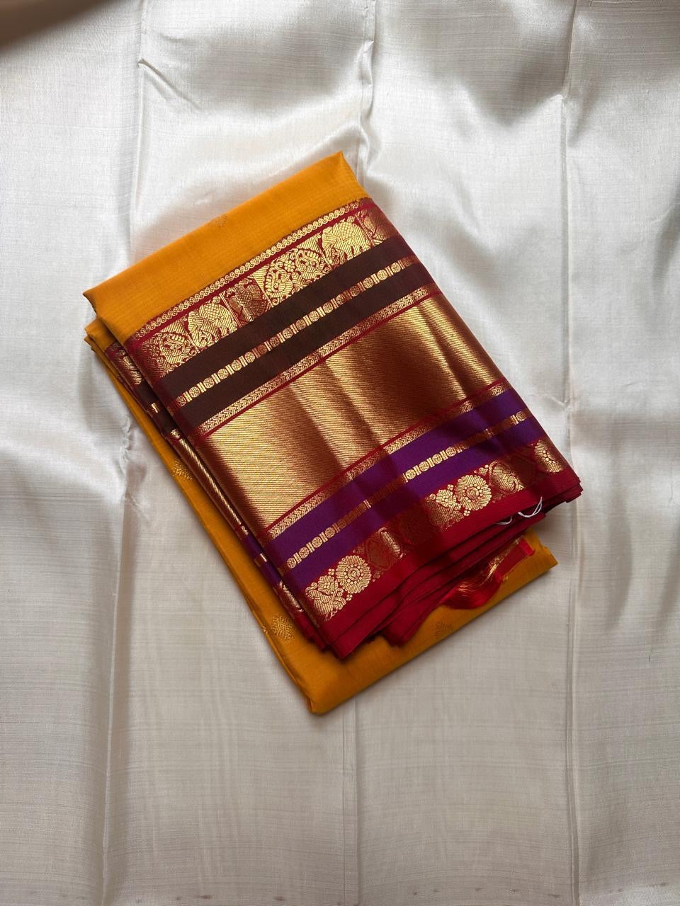 Folded Pure Kanchipuram Silk Saree in mustard yellow with wide gold zari border and maroon bands on ivory backdrop.