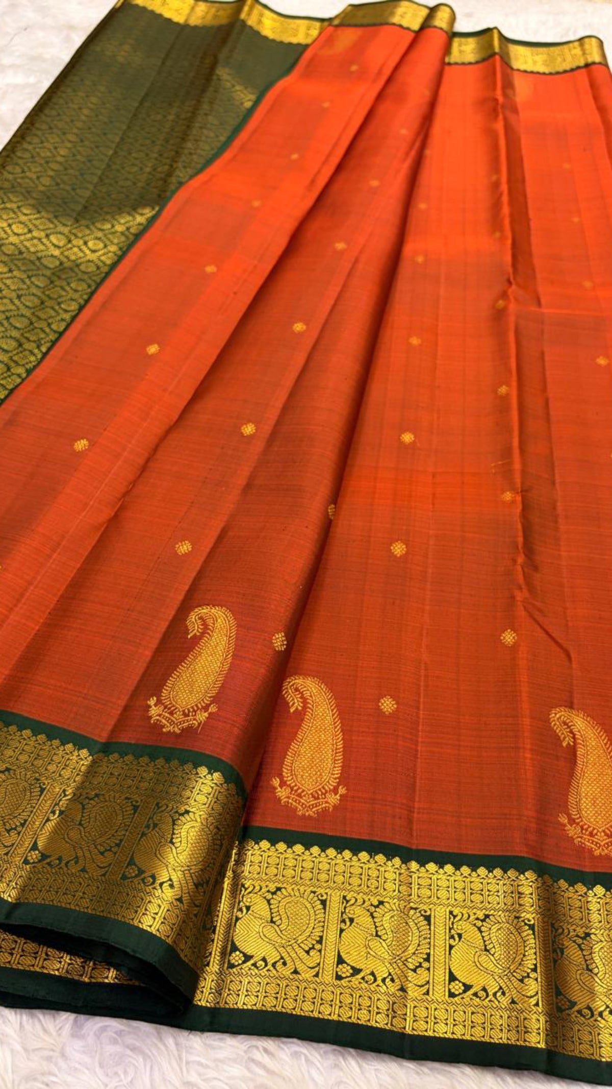 Rust orange pure silk Kanchipuram saree with small zari buttas, a bottle-green detailed paisley border, and an ornate peacock-grid kamalam pallu.