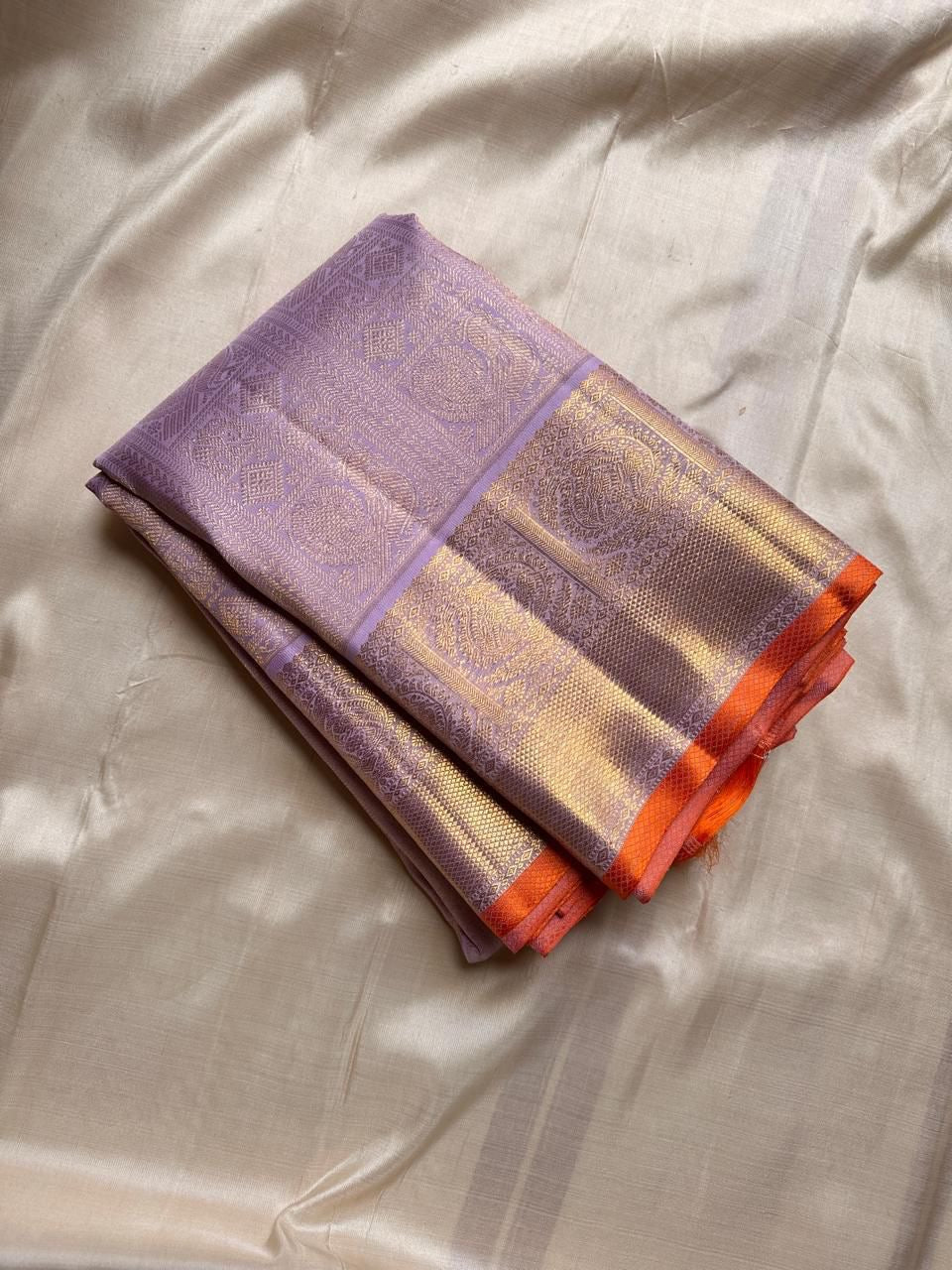 Folded lavender pure Kanchipuram silk saree with gold zari border and orange selvedge on beige silk backdrop.