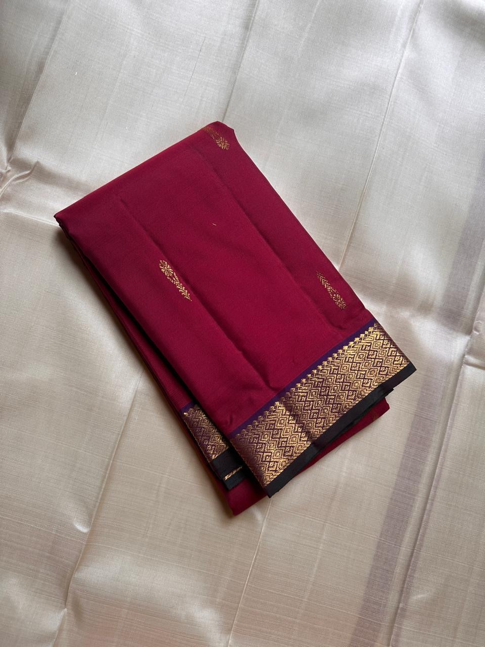 Folded deep maroon Pure Kanchipuram silk saree with small zari buttas and a gold zari woven border with dark edging.