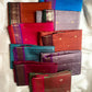 Assorted folded pure Kanchipuram silk sarees in multiple colors arranged together on a light fabric backdrop.