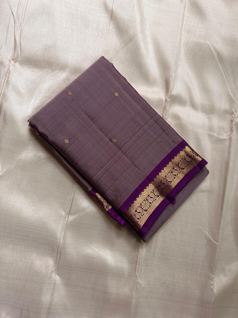 Folded mauve Kanchipuram silk saree with small gold buttas and a purple zari woven border on a light fabric backdrop.