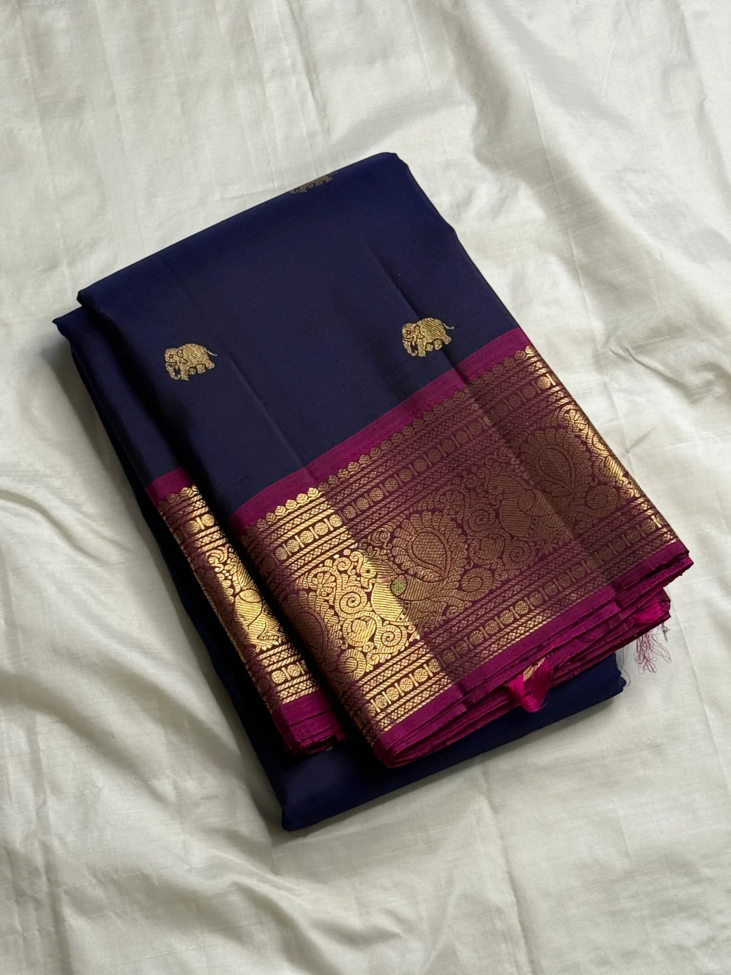 Folded navy Pure Kanchipuram silk saree with gold elephant buttas and a wide magenta-and-gold zari border on white fabric.