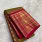 Folded olive-bronze Kanchipuram silk saree with small gold mayil buttas and a contrasting ruby-pink zari border.