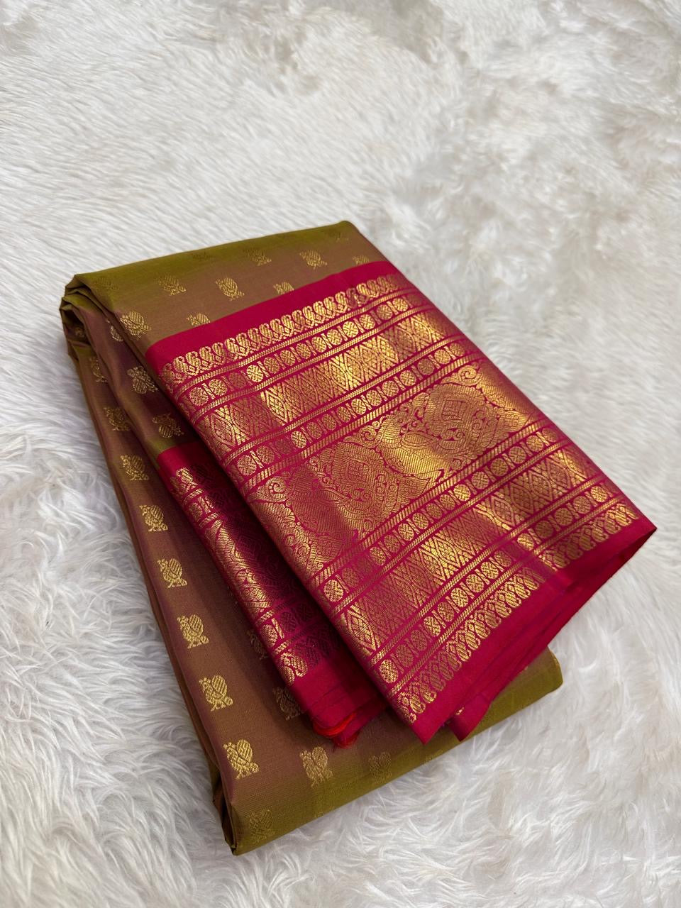 Folded olive-bronze Kanchipuram silk saree with small gold mayil buttas and a contrasting ruby-pink zari border.