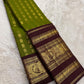 Folded olive-green Kanjivaram with gold zari bird buttas and a deep plum-brown ornamental peacock border.