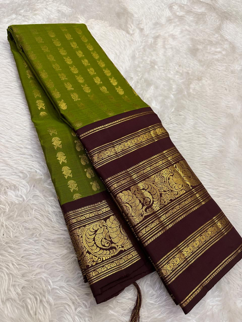 Folded olive-green Kanjivaram with gold zari bird buttas and a deep plum-brown ornamental peacock border.