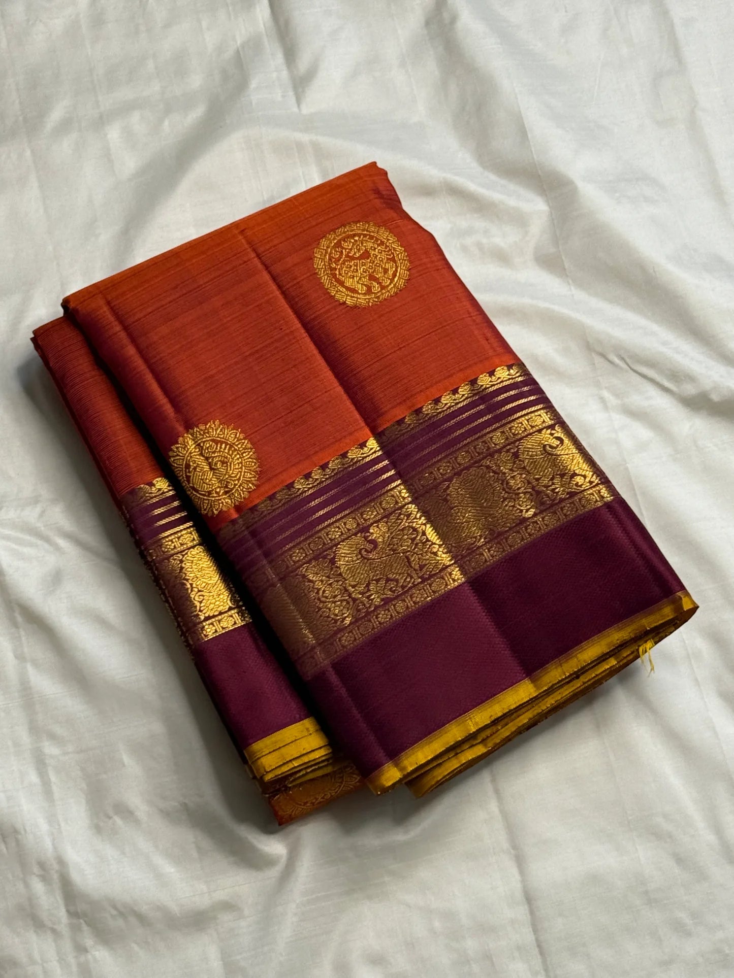 Folded rust orange Pure Kanchipuram silk saree with deep purple border and gold zari woven detailing