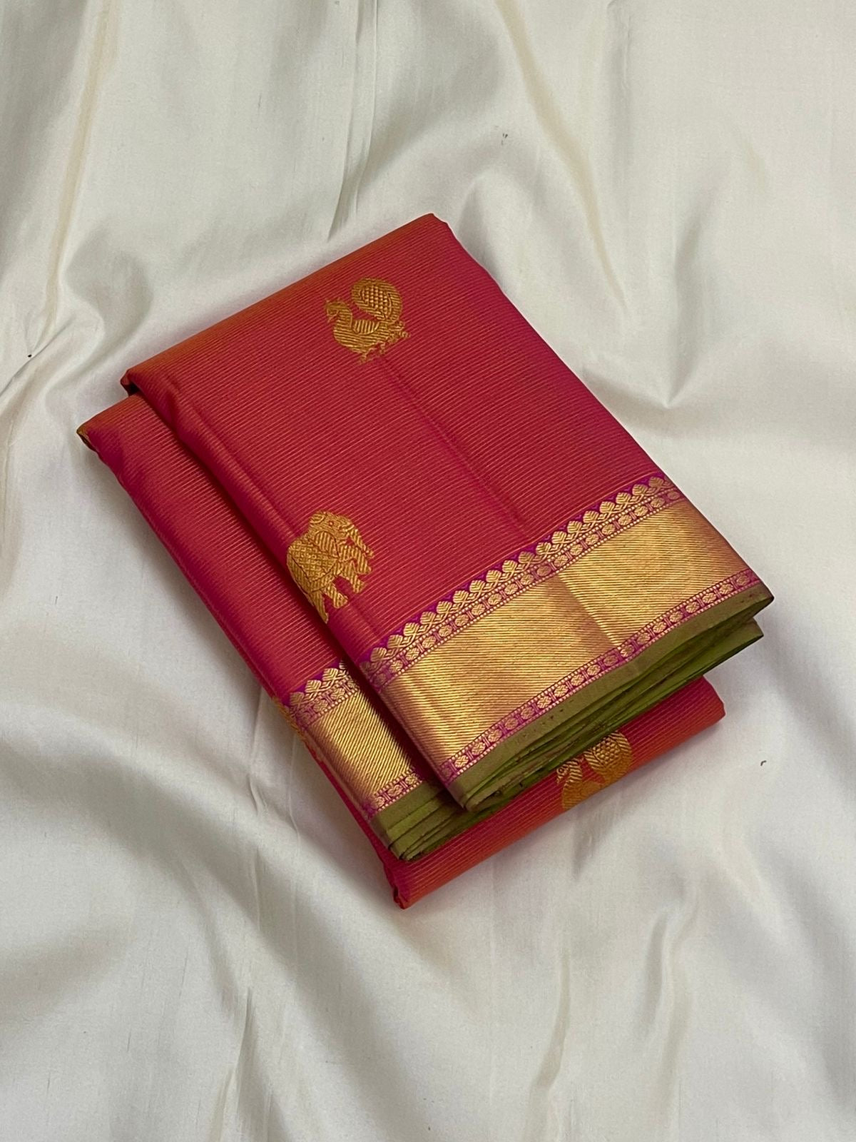 Folded rani pink vairaoosi Kanchipuram silk saree with wide gold zari border and green edge on white backdrop