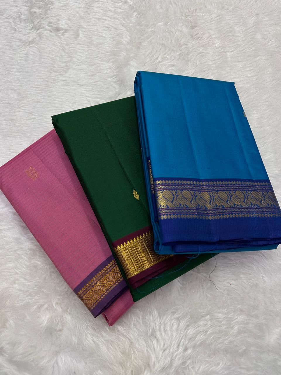Three folded silk sarees on white fur background including rose pink with purple border, green, and ink blue with zari border