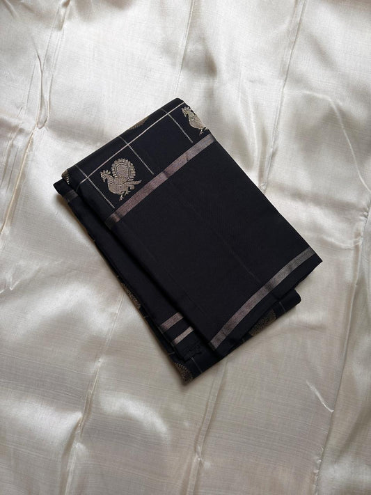 Folded black Pure Kanchipuram silk saree with zari woven check lines, peacock motif, and slim zari border.