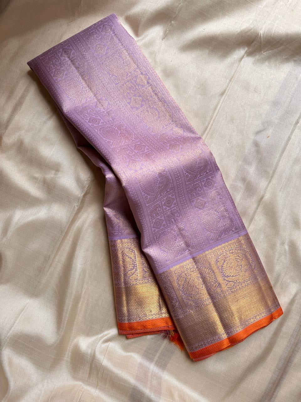 Lavender pure Kanchipuram silk saree draped to show gold zari border with orange edge on beige silk backdrop.