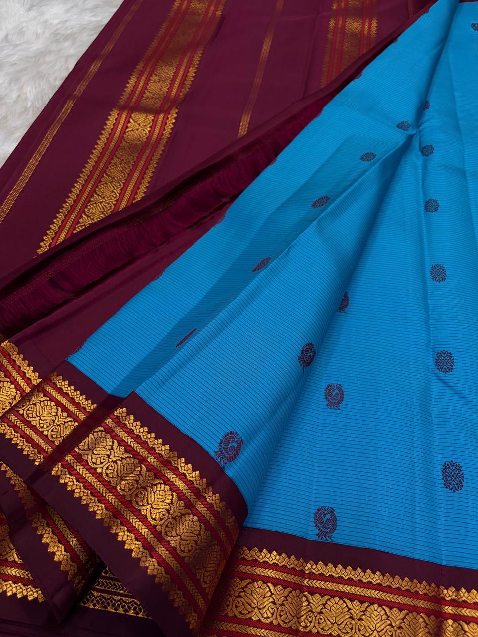 Electric peacock-blue Kanchipuram silk body showing fine Arakku hairline stripes and spaced floral silk buttas.
