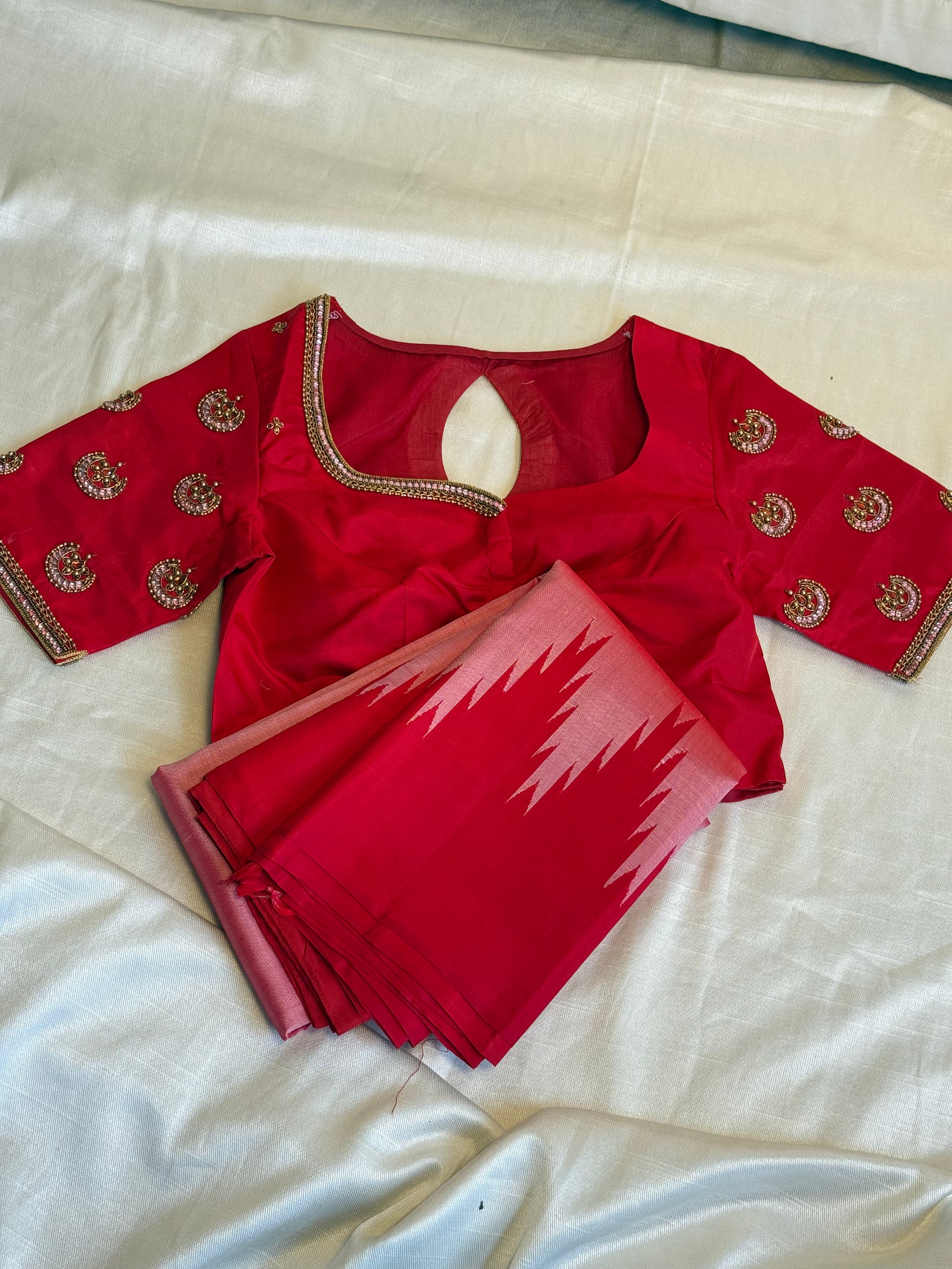 vibrant red silk saree neatly folded alongside a matching blouse featuring intricate gold and pink beadwork embroidery on the sleeves and neckline.