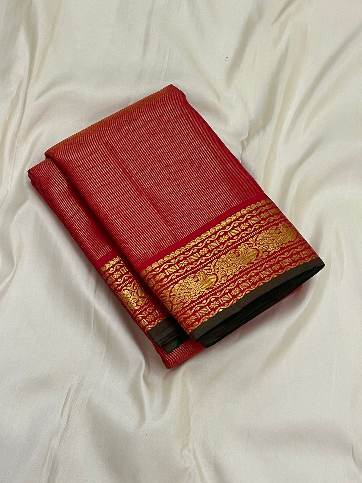 Folded vairaoosi red Kanchipuram silk saree on white fabric with gold zari border and forest green edge.