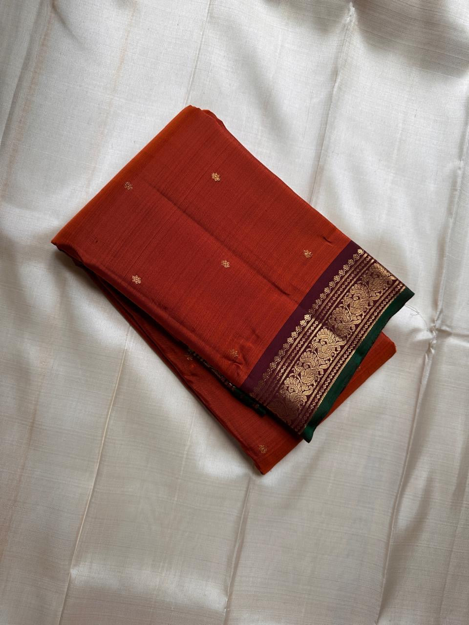 Draped rust orange Pure Kanchipuram silk saree with tiny zari buttas and a maroon zari woven border finished with a green edge.