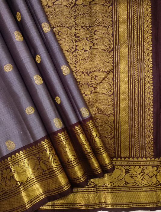 Folded slate-lavender Kanchipuram silk saree with small zari buttas and a broad Kaapi Brown peacock border