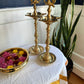 Two brass oil lamps on a table with a white cloth and a plant in the background.