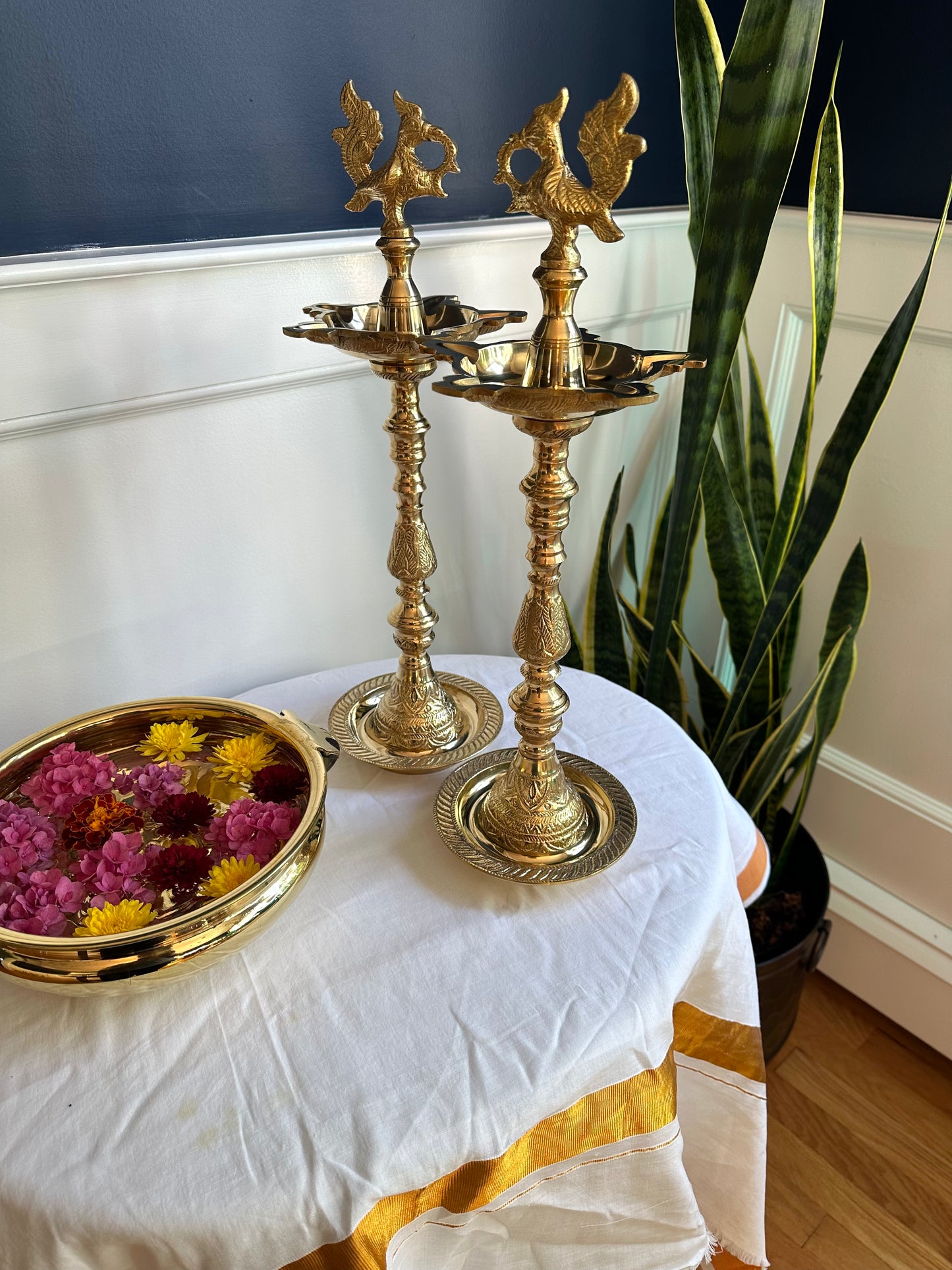 Two brass oil lamps on a table with a white cloth and a plant in the background.